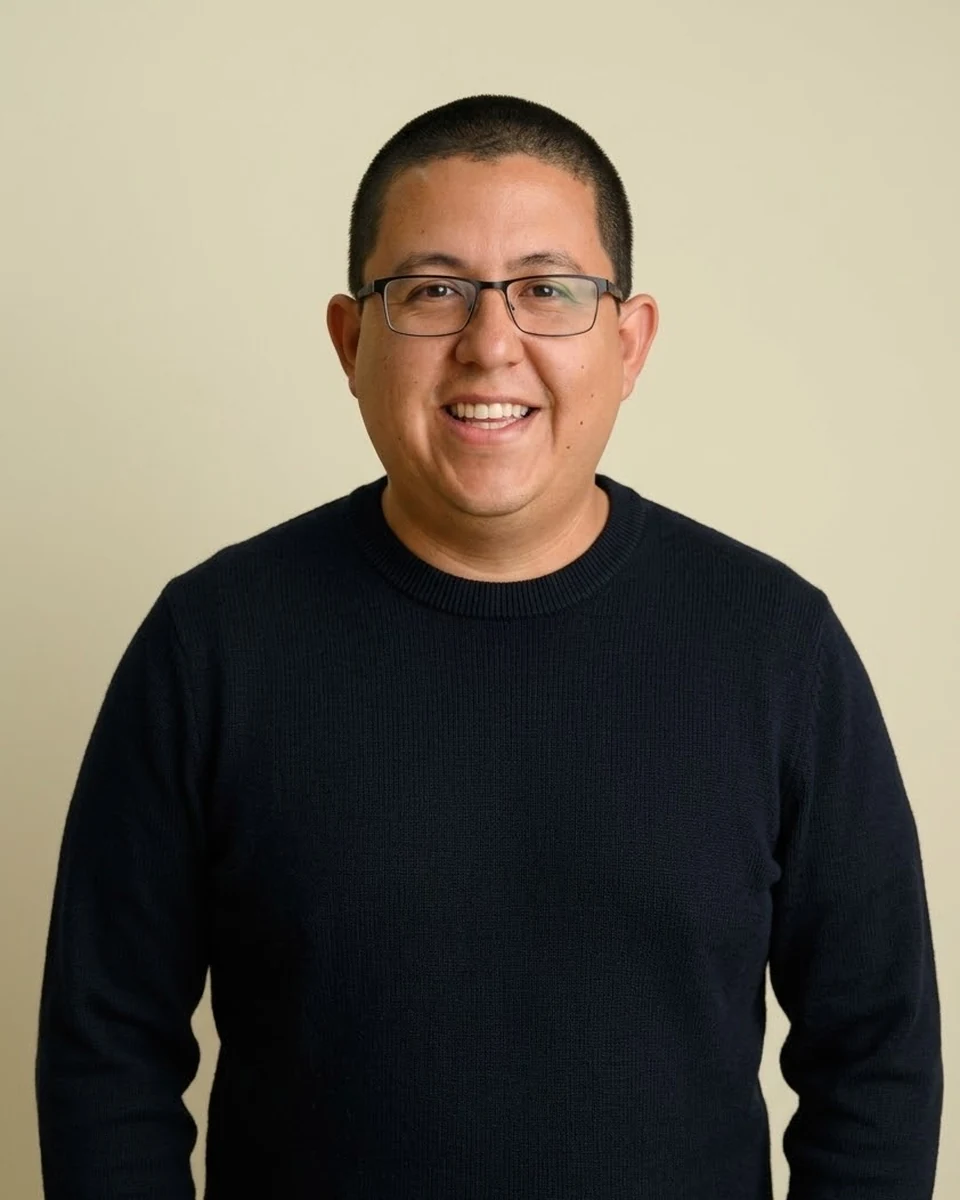 Cristian Penarrieta: dark crew-neck sweater, rectangular glasses, looking straight at the camera against a warm off-white wall.